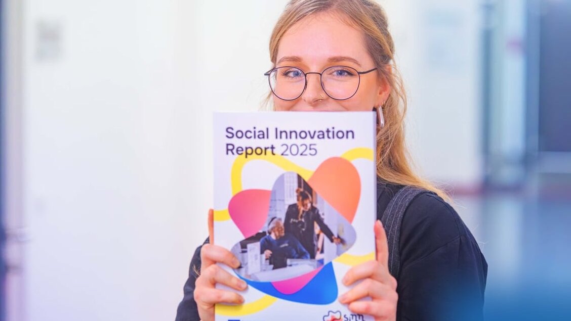 Eine Frau ein Heft hoch und zeigt es in die Kamera. Auf dem Heft steht Social Innovation Report 2025. - Social Innovation Report 2025 - Werft einen Blick rein