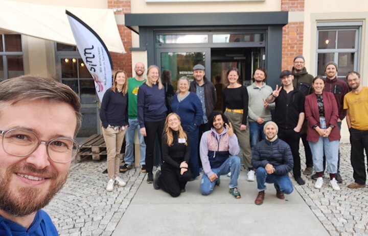 Im Hintergrund steht eine Gruppe an menschen und lächelt in die Hintergrund. Links Vorne ist ein Gesicht von einem Mann zu sehen. Auch er lächelt in die Kamera und macht das Foto. - Link: Rückblick: 2 Jahre SINNkubator