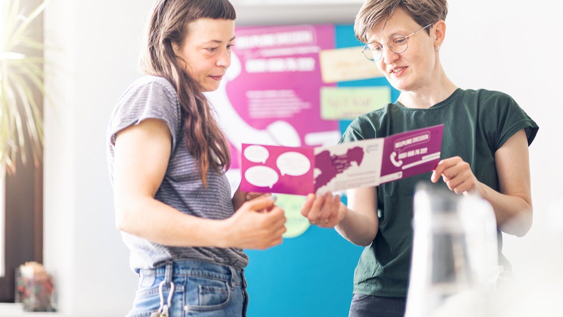 Zwei Frauen stehen nebeneinander. Sie schauen gemeinsam in eine Broschüre. - Das Innovations-Sofa. Diesmal mit Helpline Sachsen