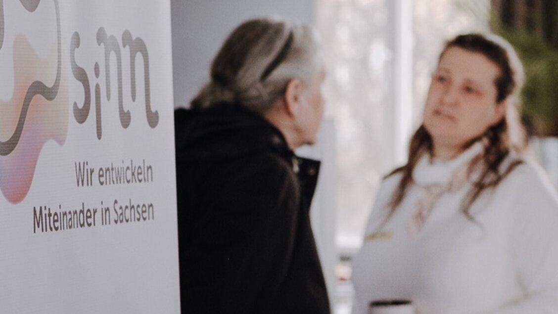 Zwei Personen sprechen miteinander. Das Bild ist unscharf. - Nehmt teil an unserer Bürger:innenbefragung!