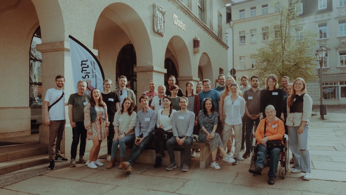 SINNkubator Kick-Off April 2024 Über 20 Menschen stehen und sitzen eng als große Gruppe und schauen in die Kamera. Dahinter ist ein Gebäude und ein Baum. - Die erste Runde des SINNkubator-Programms ist gestartet