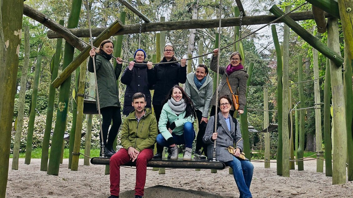 Acht Menschen sitzen und stehen auf einer riesigen Schaukel aus Holz. - Das Innovations-Sofa. Diesmal im Interview: das Team von Kraft-Copilot