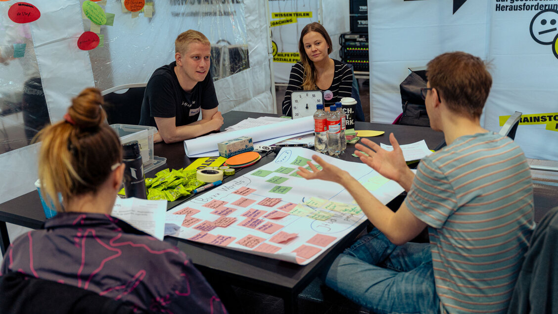 Vier Menschen sitzen an einem Tisch und reden miteinander. Vor ihnen liegen Blätter, Zettel und Stifte. - Neue SINN Events SINN Werkstatt in Mitweida und KI Workshop-Reihe