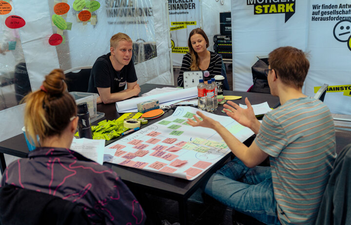 Vier Menschen sitzen an einem Tisch und reden miteinander. Vor ihnen liegen Blätter, Zettel und Stifte. - Link: Neue SINN Events SINN Werkstatt in Mitweida und KI Workshop-Reihe