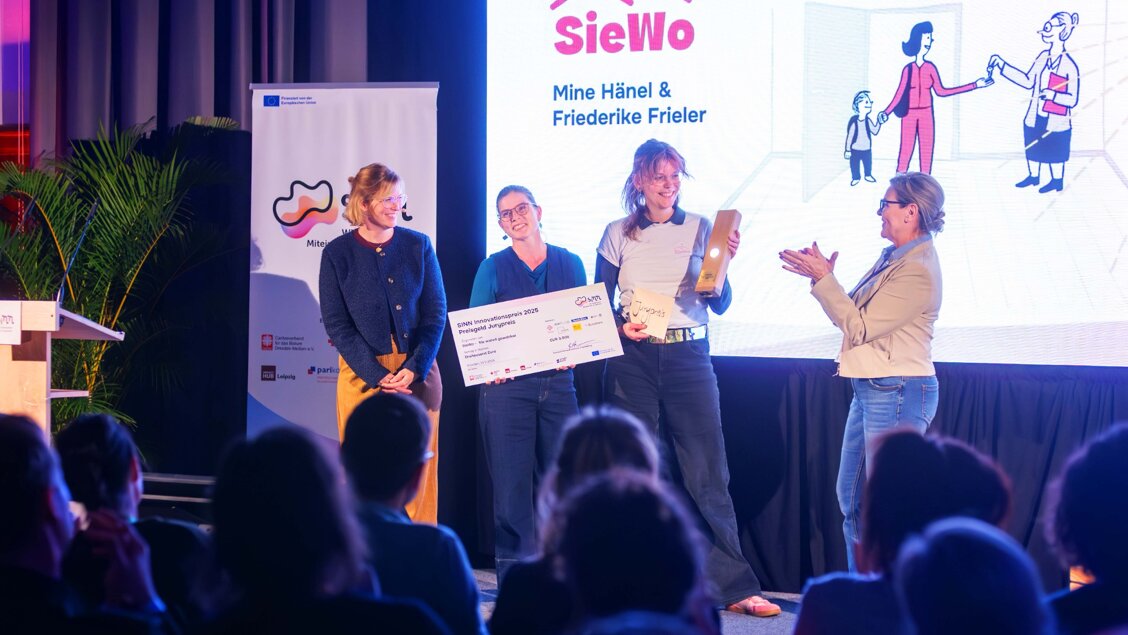 Vier Menschen stehen auf der Bühne. Zwei davon bekommen einen Preis überreicht. Man sieht die Köpfe des Publikums von hinten. - Ihr könnt nur noch bis zum 08.10. abstimmen: SINN Innovationspreis