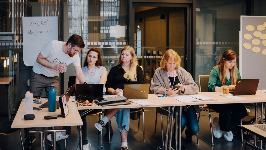 Vier Menschen sitzen an einem Laptop. Eine Person steht und erklärt zwei der sitzenden Menschen etwas. - Geballtes Wissen: Online-Fachtag zu Digitalisierung am 21.08.