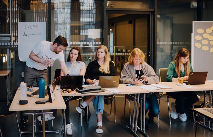 Vier Menschen sitzen an einem Laptop. Eine Person steht und erklärt zwei der sitzenden Menschen etwas. - Link: Geballtes Wissen: Online-Fachtag zu Digitalisierung am 21.08.