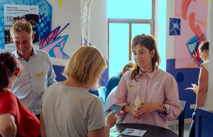Verschiedene Menschen stehen in einem Raum. Sie reden miteinander und lächeln sich an. - Link: Menschen im Austausch - Roadshow Riesa am 28.08.