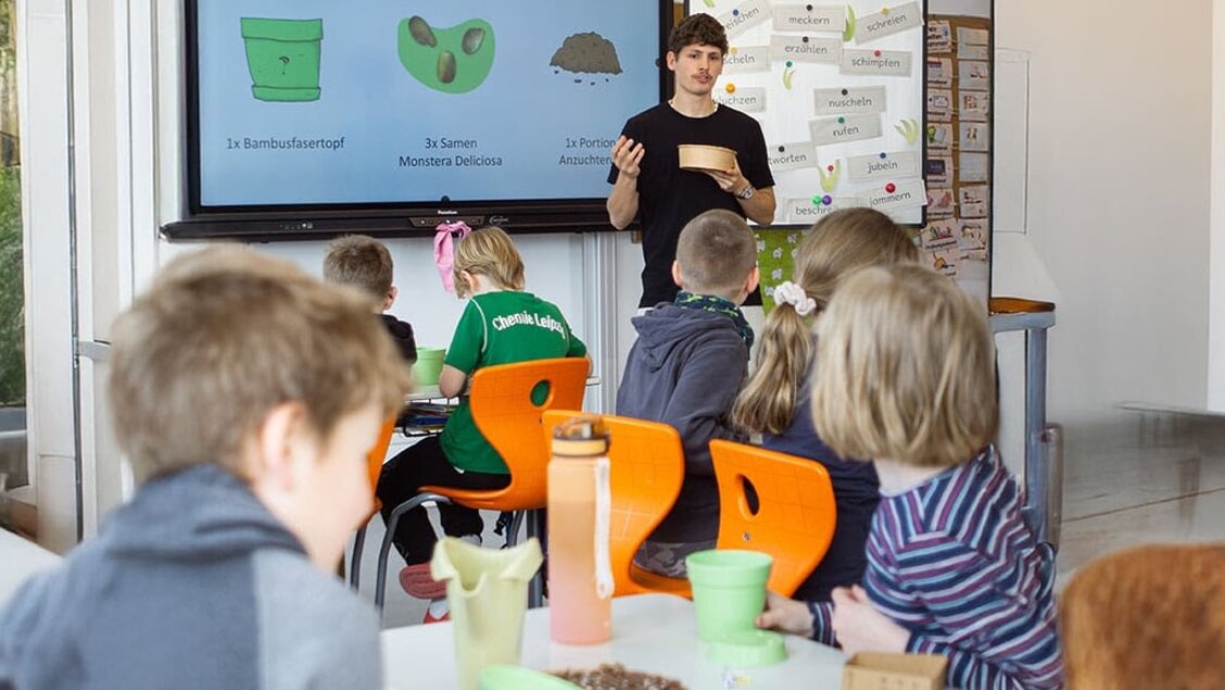 Ein Mann steht an einer Tafel und erklärt Kindern etwas. Die Kinder sitzen und sind nur von hinten zu sehen. - Das Innovations-Sofa. Diesmal mit Pflanzpaket