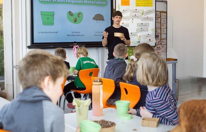Ein Mann steht an einer Tafel und erklärt Kindern etwas. Die Kinder sitzen und sind nur von hinten zu sehen. - Link: Das Innovations-Sofa. Diesmal mit Pflanzpaket