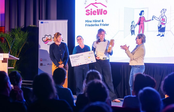 Vier Personen stehen auf der Bühne. Zwei von ihnen bekommen einen Preis und einen Scheck gereicht. - Link: SINNovationsfest - Publikum, Präsentationen, Preise