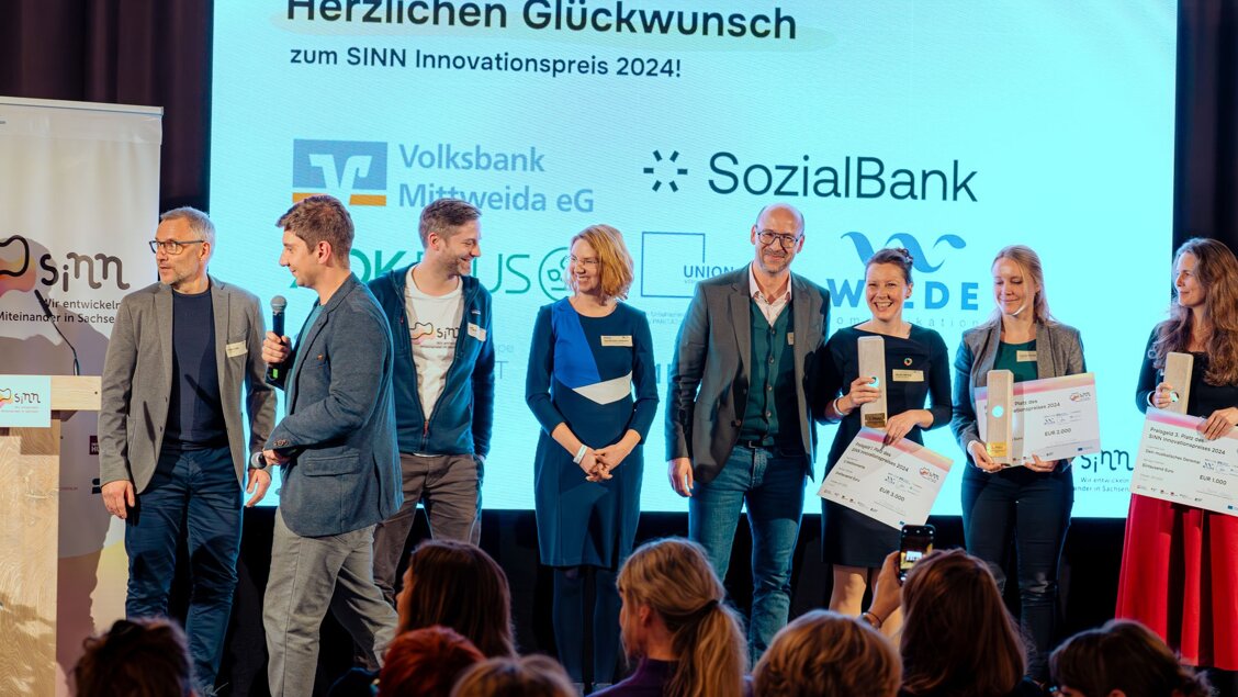 Mehrere Menschen stehen auf einer Bühne. Sie sind in Bewegung und lächeln. - Abstimmen für den SINN Innovationspreis und Ticket für die Preisvergabe sichern