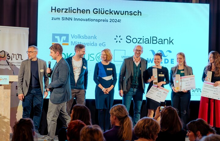 Mehrere Menschen stehen auf einer Bühne. Sie sind in Bewegung und lächeln. - Link: Abstimmen für den SINN Innovationspreis und Ticket für die Preisvergabe sichern