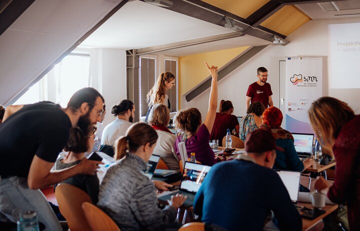 Viele Menschen sitzen an Tischen und arbeiten. Eine Person hebt die Hand, um etwas zu fragen. - Link: Noch bis zum 22.03. für das SINNkubator-Programm bewerben