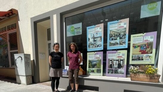 Zwei Frauen stehen vor einem Haus. Das haus hat eine großes Schaufenster in dem bunte Plakate zu sehen sind. - Wanderausstellung zu sozialen Innovationen zieht weiter nach Mittweida