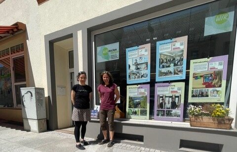 Zwei Frauen stehen vor einem Haus. Das haus hat eine großes Schaufenster in dem bunte Plakate zu sehen sind. - Link: Wanderausstellung zu sozialen Innovationen zieht weiter nach Mittweida