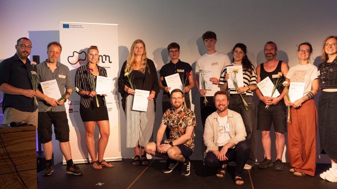 Viele Menschen stehen und sitzen. Sie lächeln in die Kamera. Manche haben eine Blume und urkunde in der Hand. - Innovationsbühne DD - SINNkubator-Finale in groß