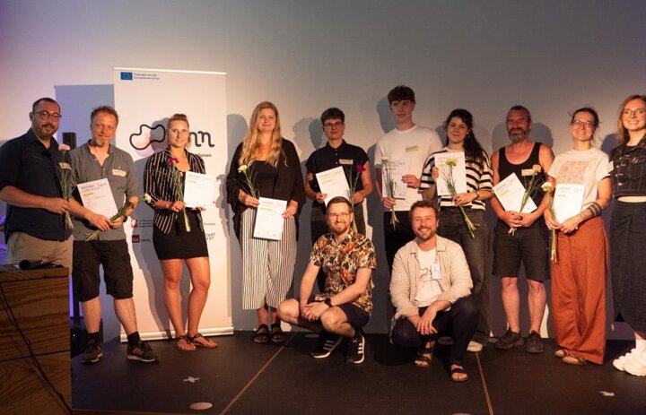 Viele Menschen stehen und sitzen. Sie lächeln in die Kamera. Manche haben eine Blume und urkunde in der Hand. - Link: Innovationsbühne DD - SINNkubator-Finale in groß